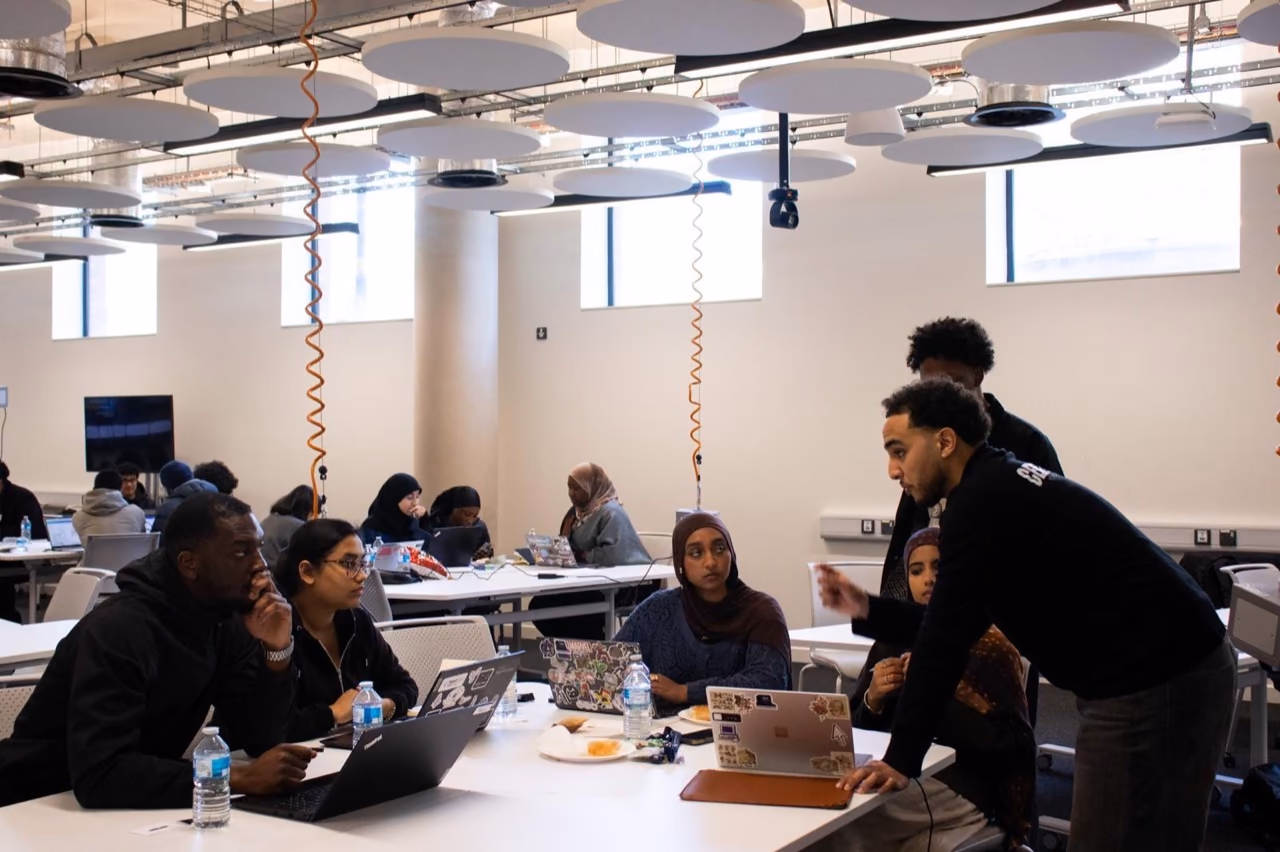 Hamza leaning over laptops, working with a team at a hackathon